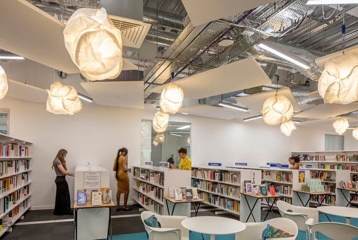 People checking out books at London Academy of Excellence's library, well-lit library with white walls, unique hanging light fixtures, and exposed ceiling pipes.