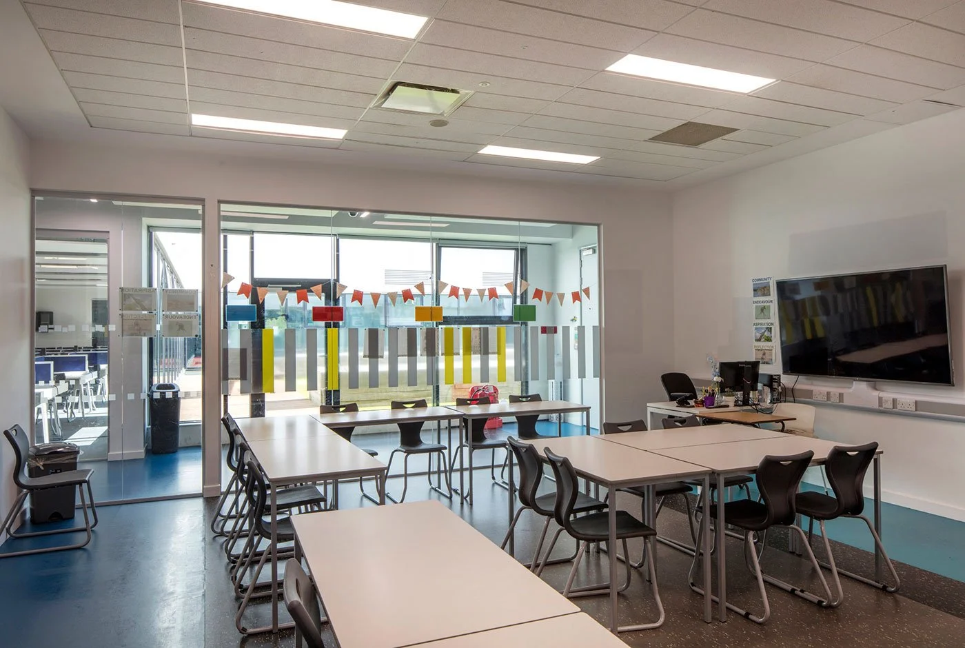 Empty classroom with tables, chairs, a large TV, and colorful decorations on the windows.