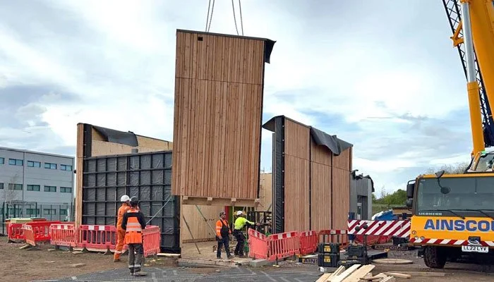 Workers wearing orange safety vests and helmets are constructing a building with wooden and black exterior panels, with a crane on the right side lifting a large wooden panel, surrounded by orange safety barriers outdoors on a cloudy day.
