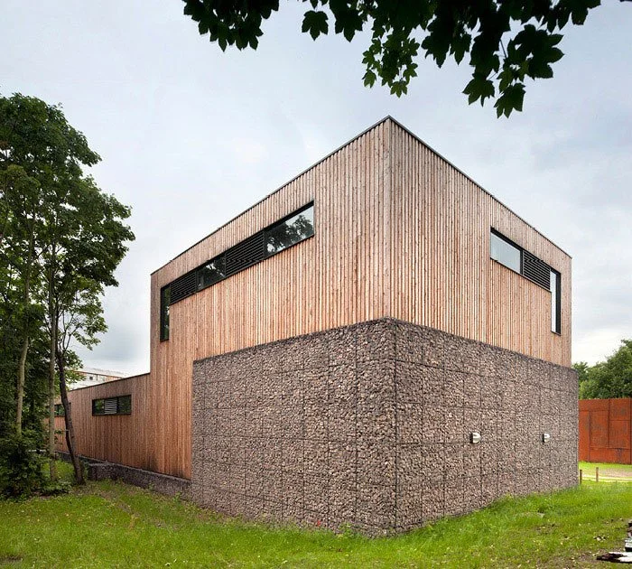 Modern two-story house with a wooden upper section and a textured concrete lower section, surrounded by green grass and trees.