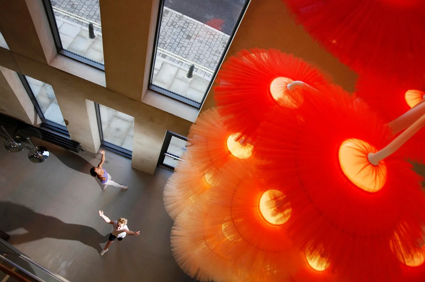 Looking up at a beige interior with large windows and two people dancing on the floor, with a bright orange chandelier hanging from the ceiling.