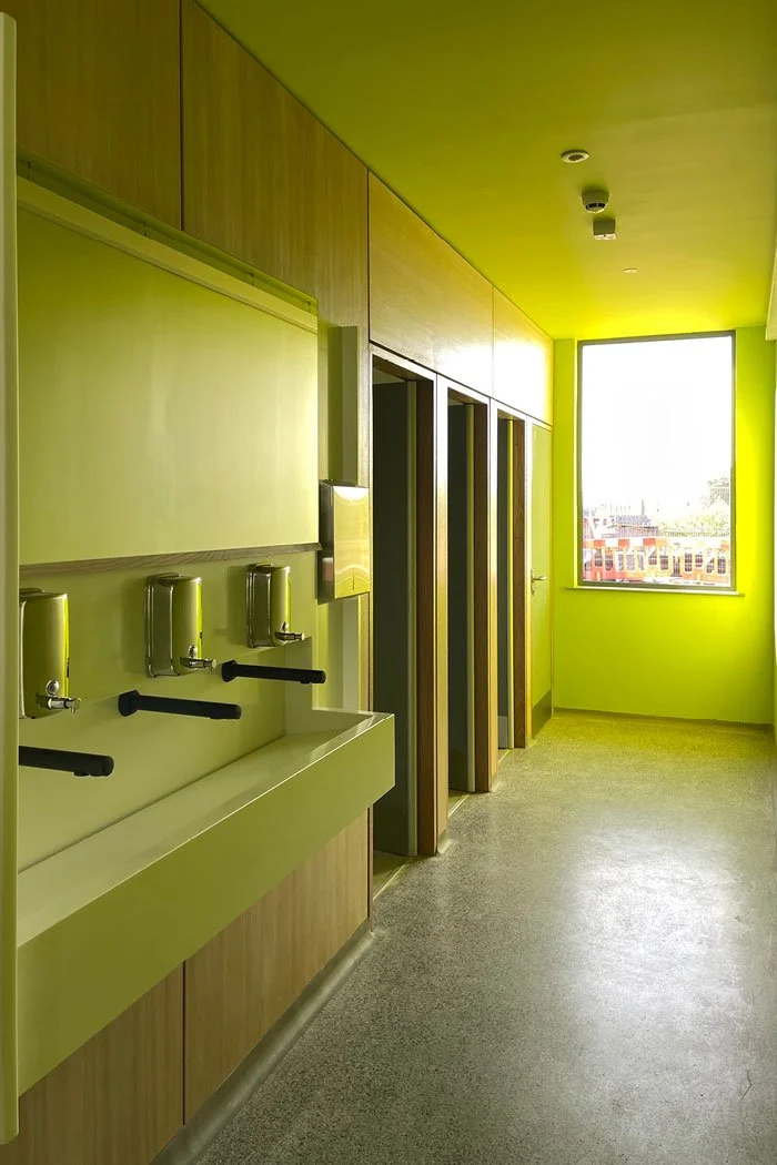 A hallway with a lime green wall, a sink with three soap dispensers, and multiple bathroom stalls. There is a large window at the end of the hallway letting in natural light.