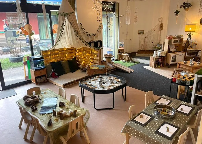 A cozy indoor space decorated with a string of lights, pillows, and a canopy, with a large window showing the street outside. There are tables set for a meal, a play area with toys and books, and various shelves and storage units filled with children's items.