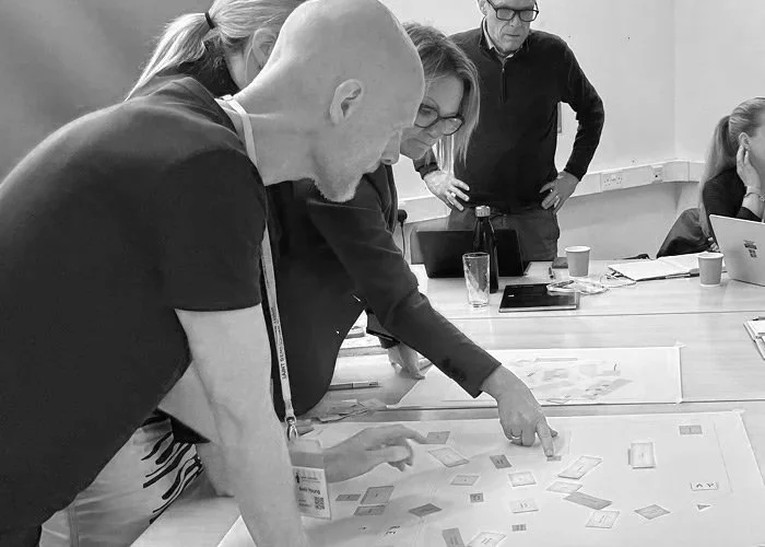 Group of people gathered around a table, analyzing and discussing materials spread out on the table in a meeting or collaborative setting.