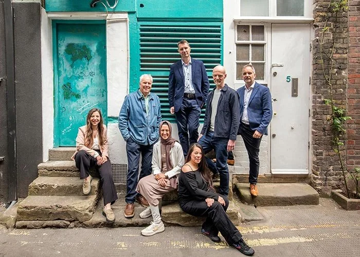 The LBY Architects Team, a group of seven people standing and sitting – posing to the camera – on stairs outside a teal-coloured building with a white door and a vent.