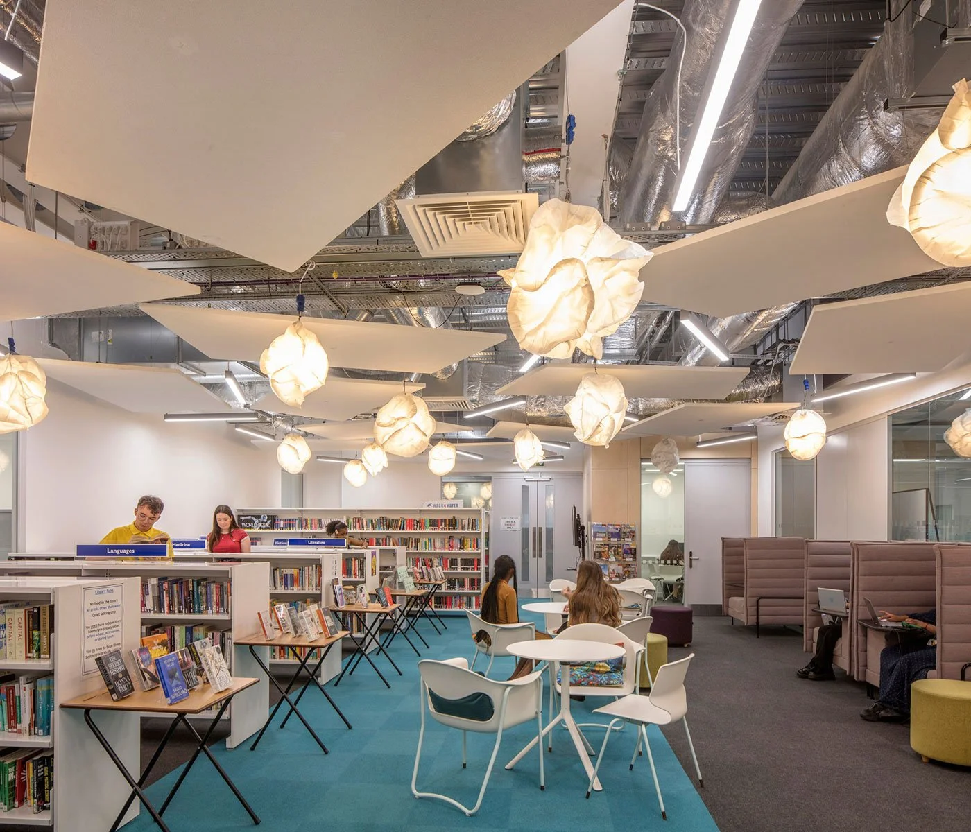 Interior of a modern library with people reading and browsing books, illuminated by decorative hanging lamps and "Languages" and "Literature" sections in the background.