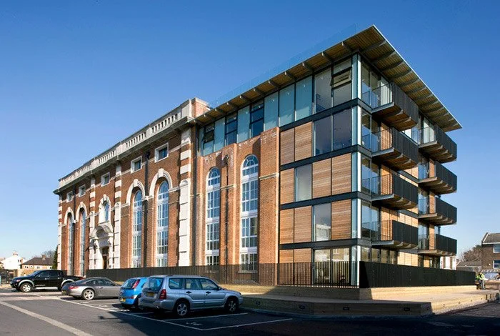 A modern multi-story building with large glass windows and balconies, adjacent to a historic brick building with arched windows and detailed stonework, with parked cars in front.