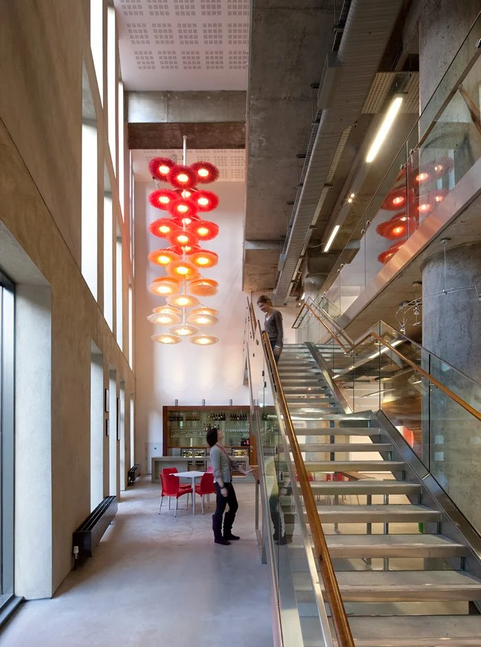 Interior of a modern building with a staircase leading to an upper level, large windows, colorful hanging lights, and two people, one ascending and one standing near the staircase.