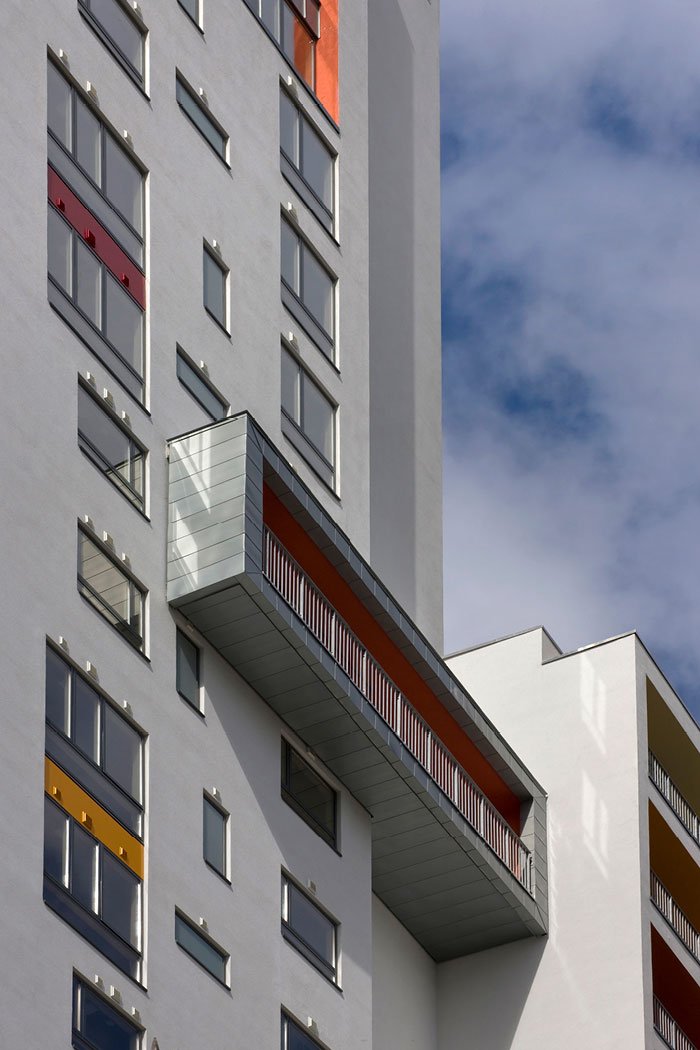 Modern apartment building with white exterior, various colorful window frames, and a protruding balcony with orange and gray details, set against a blue sky with clouds.