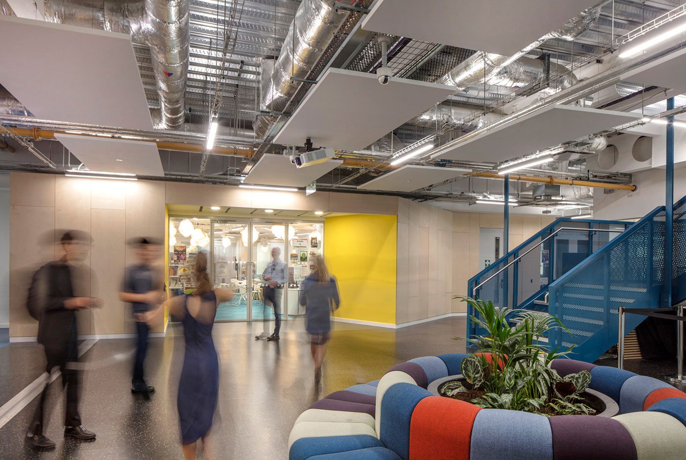 People walking in a modern office or coworking space with colorful furniture, an indoor plant, and a staircase, under exposed ceiling pipes and panels.