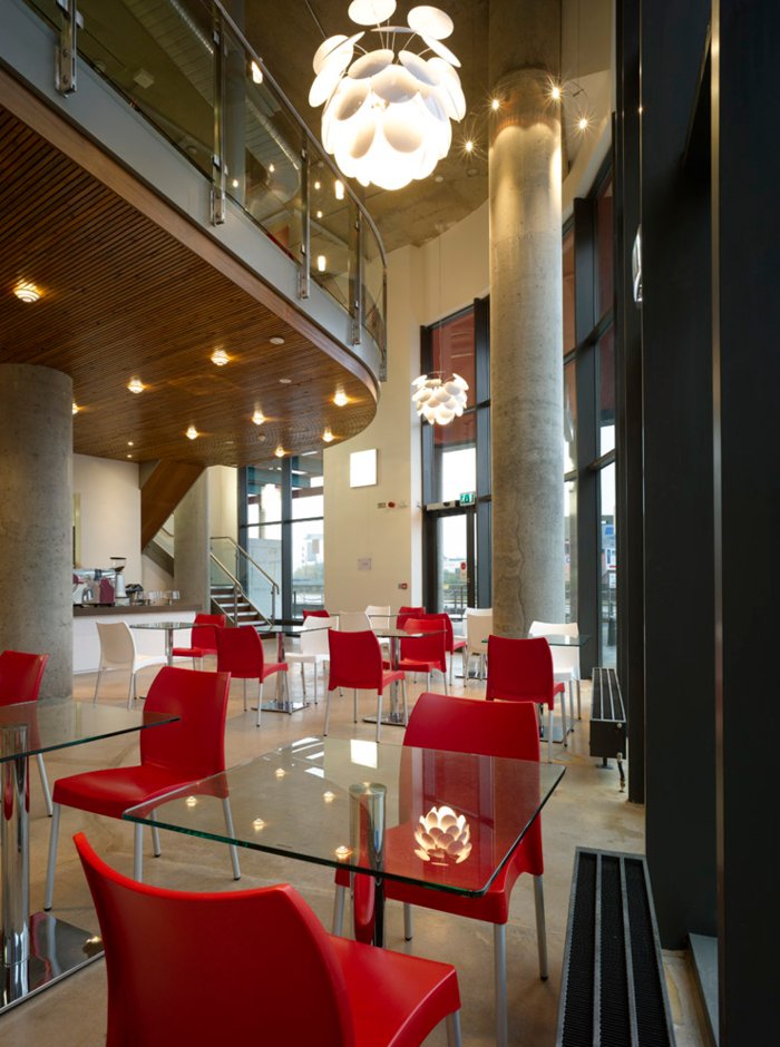 Modern cafe or restaurant interior with red and white chairs around glass tables, large windows, polished concrete floors, and contemporary chandeliers, featuring a mezzanine level with glass railing and wooden ceiling accents.