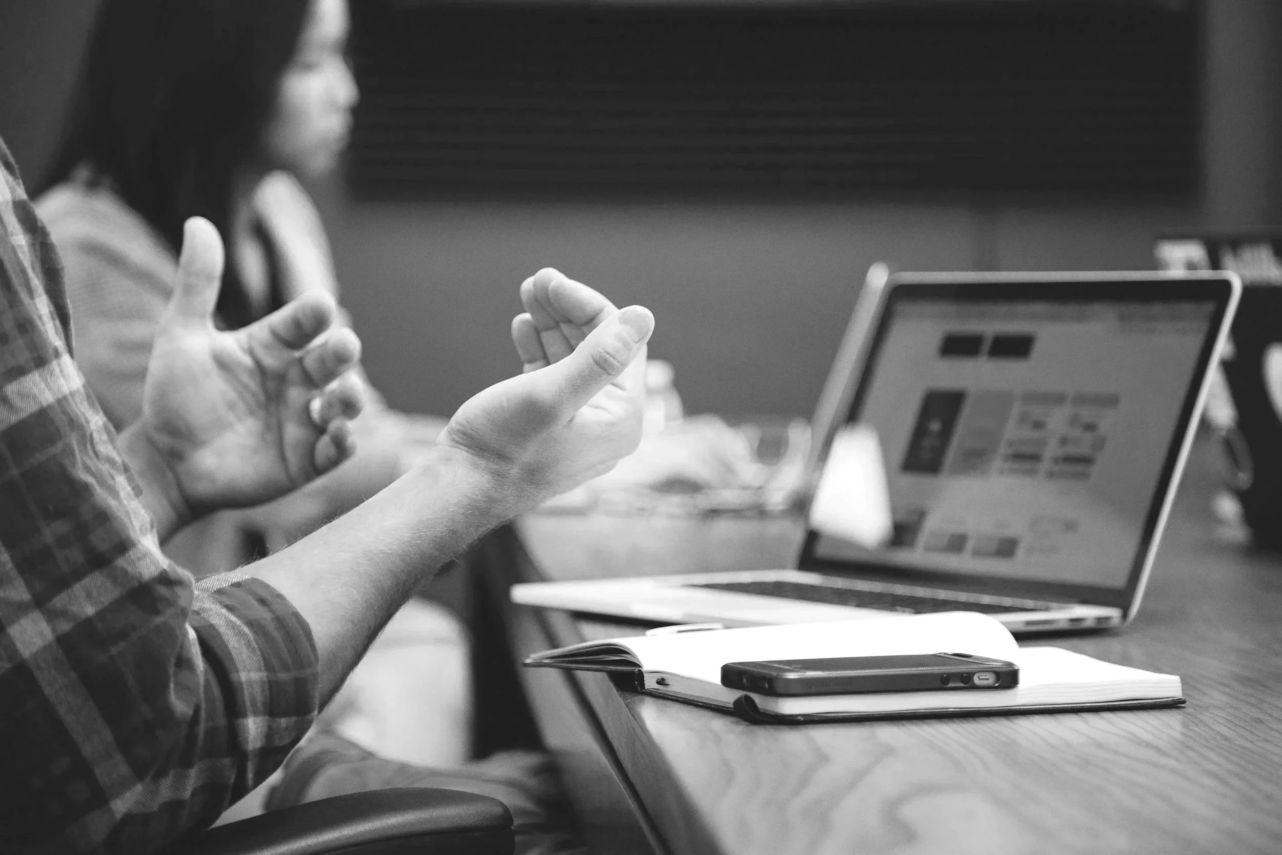 People sitting at a desk with a laptop, smartphone, and notebook engaged in a discussion.