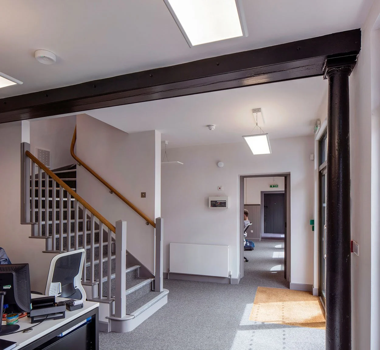 Interior view of a building lobby or reception area with a staircase on the left, ceiling lights, and an open door leading to another room with a person sitting at a desk.