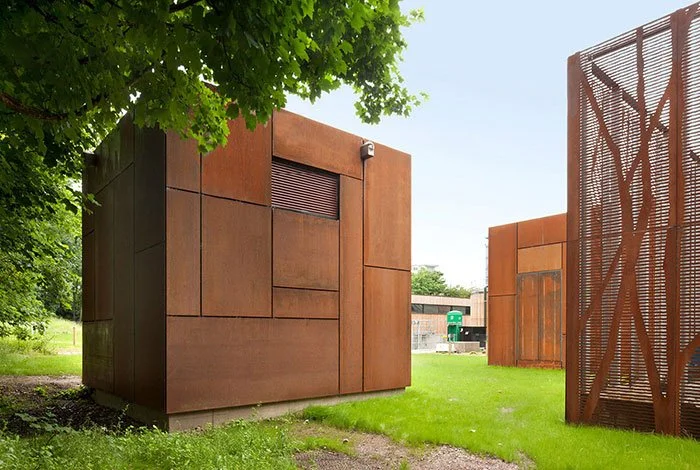 Modern rust-colored metal structures with geometric designs in a park-like setting with green grass and trees.