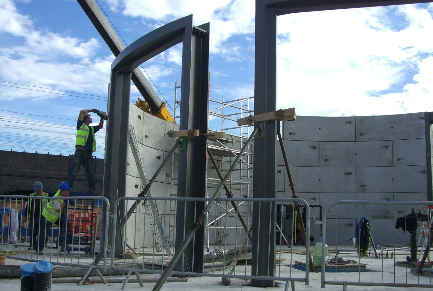 Construction workers building a concrete wall and steel framework on a bright day with blue sky and clouds.