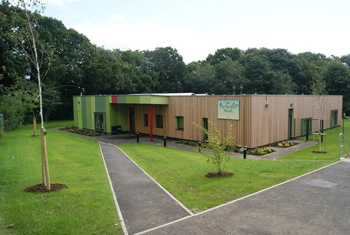 A full external view of the Enchanted Wood Nursery, a single-story building with a combination of green, red, and wood exterior, surrounded by a lawn and trees, with paved walkways and a small playground area.