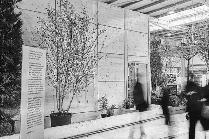 Indoor scene with people walking past a concrete wall, potted plants, a QR code, and informational signage.