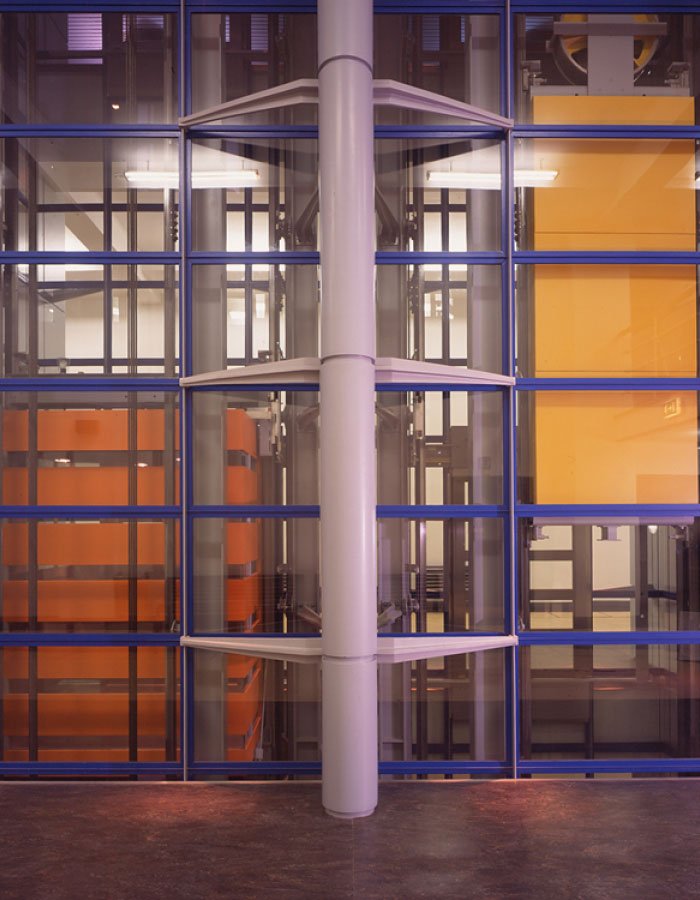 Colourful internal workings of the Science Museum elevator with visible internal mechanisms through the glass walls, displaying orange and yellow panels against the blue frame panels, with a large grey support beam central to the elevator shaft.