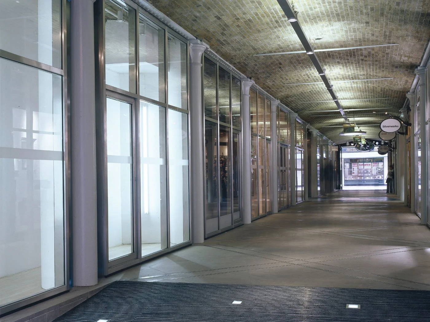 A view down the empty shopping mall corridor of Thomas Neal’s Centre with glass storefronts, tile flooring, and ceiling with track lighting, at dusk.