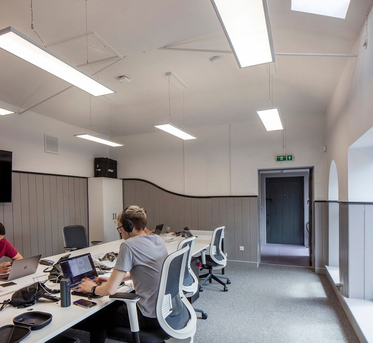 Office meeting room with people working at a long white table, using laptops and wearing headphones, with gray walls, ceiling lights, and a door in the background.