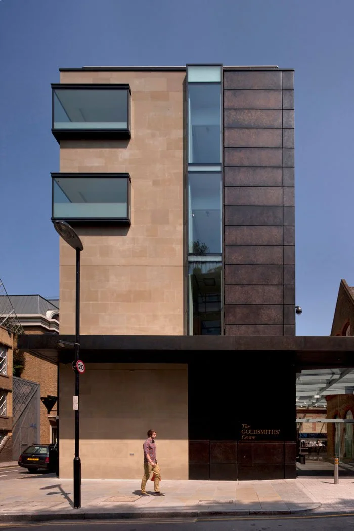 Modern multi-story building with beige and dark brown exterior walls, glass balconies, and large glass windows, with a person walking on the sidewalk in front.