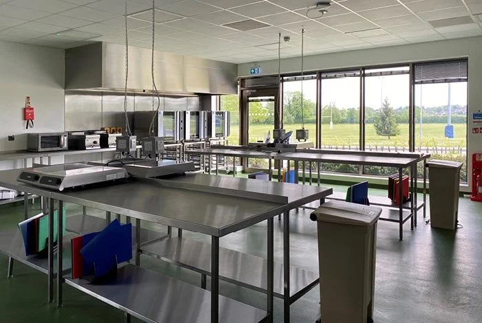 Empty commercial kitchen or cafeteria with stainless steel tables, large windows overlooking a grassy field, and various appliances and equipment.