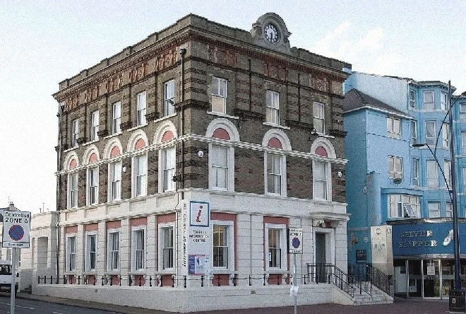Historic multi-story building of Tourist Information Centre at Great Yarmouth with a clock on top, located on a street corner, with neighbouring blue building and various street signs in a small town setting.