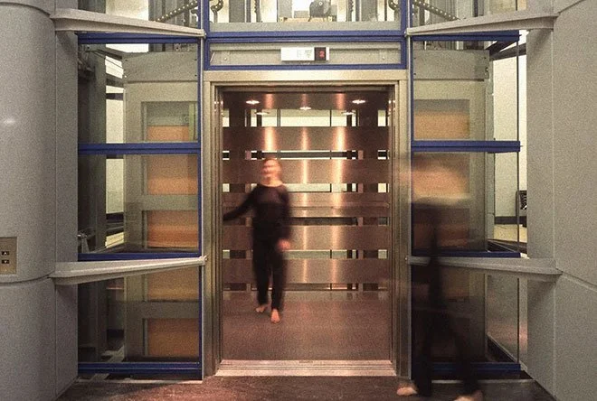 A person is walking out of the Science Museum elevator with a metallic door, some people are blurry in motion, and there's a woman on the left side entering the elevator.