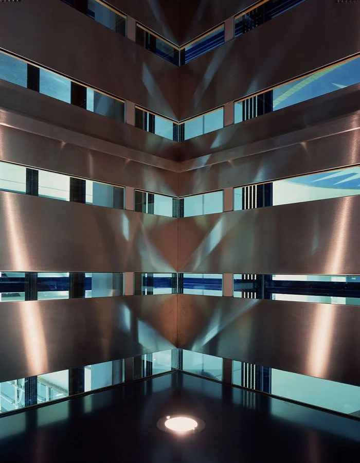 A stylishly lite shot of the corner of the Science Museum building interior looking out of one corner, with angular metal walls and large glass windows reflecting sunlight through narrow gaps in the wall panels.