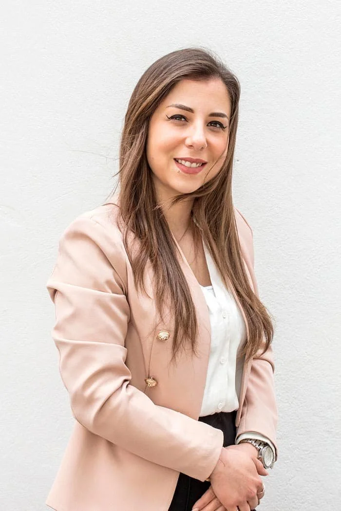 A woman with long brown hair, light skin, and a piercing just below her lower lip, smiling and standing against a plain white wall. She is wearing a light pink blazer over a white blouse, with a watch on her left wrist.