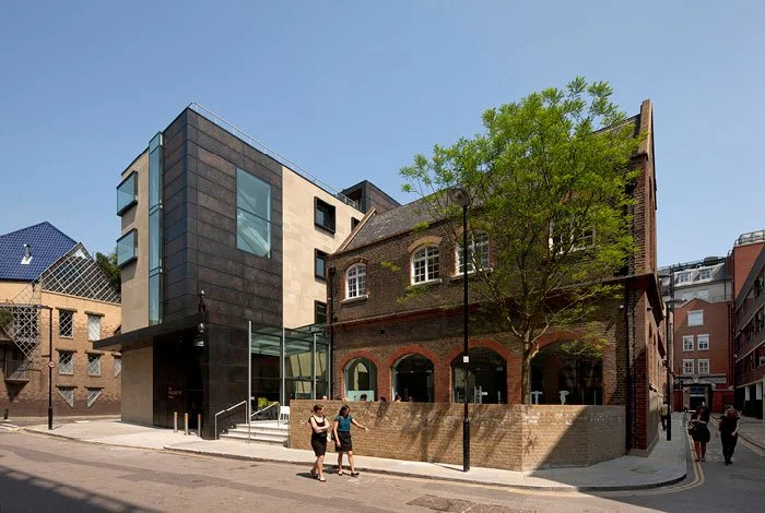 A modern building with dark and beige exterior walls and large windows, next to an older brick building with arched windows and a tree, on a city street with pedestrians.