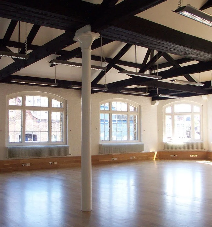 An empty, spacious room inside Thomas Neal’s Centre with large arched windows, a white pillar in the centre, and a black ceiling with exposed original wooden beams.