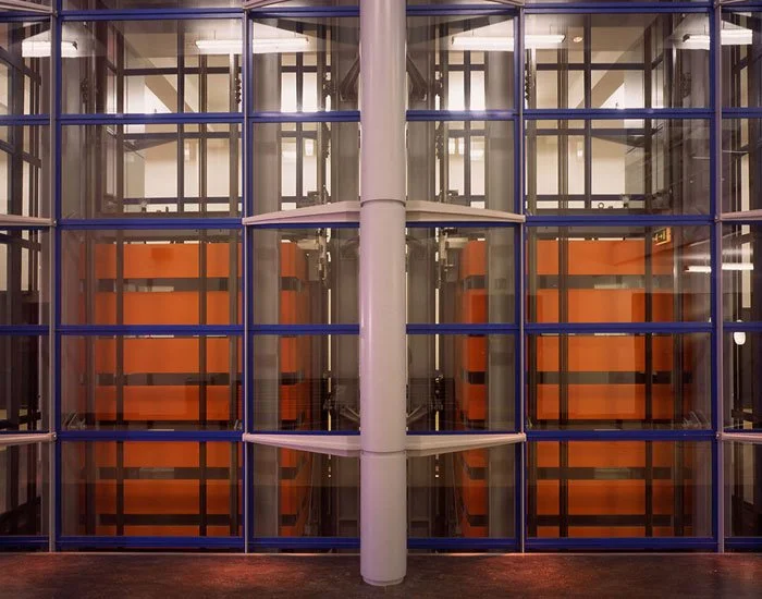 Colourful internal workings of the Science Museum elevator with visible internal mechanisms through the glass walls, displaying symmetrical orange panels against the blue frame panels, with a large grey support beam central to the elevator shaft.