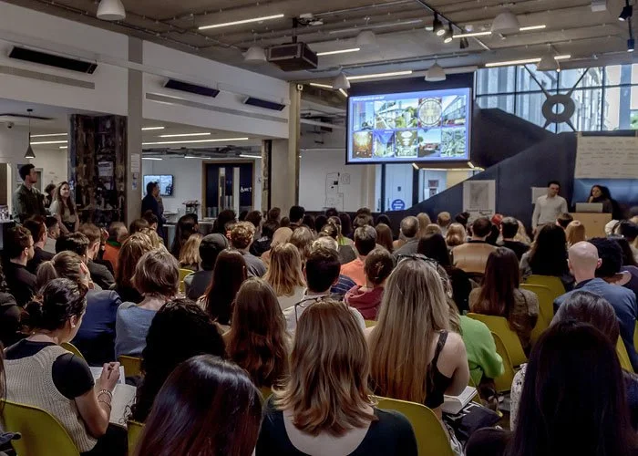 Large group of people attending a presentation or lecture in a modern conference room with a large screen displaying images at the front.