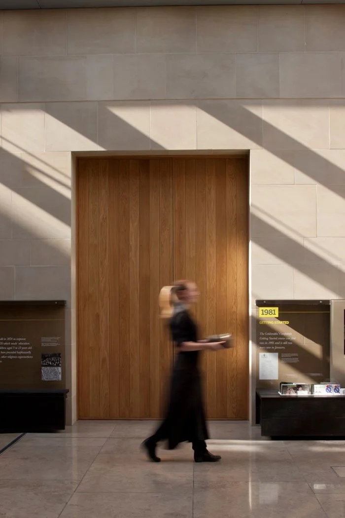 A person with blonde hair wearing a black dress and black shoes walking in front of a large wooden door in a well-lit indoor space, with informational display panels on either side of the door.
