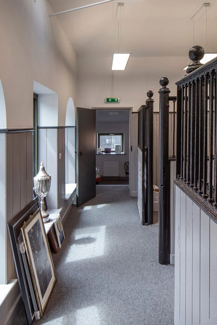 Interior view of a hallway of Mascalls Stable Block with grey carpet, white walls, and windows on the left side. Framed pictures lean against the wall, a silver trophy on display. An open door leads to another room with a window.