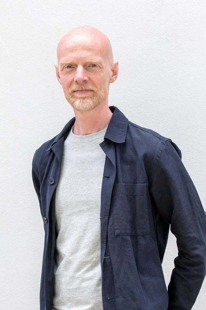 A bald man with a light beard and mustache wearing a gray t-shirt and a blue jacket, standing against a plain white wall.