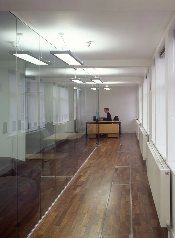 The empty office corridor inside Silver House with a glass wall on the left, a wooden floor, and a man working at a desk in the far background.