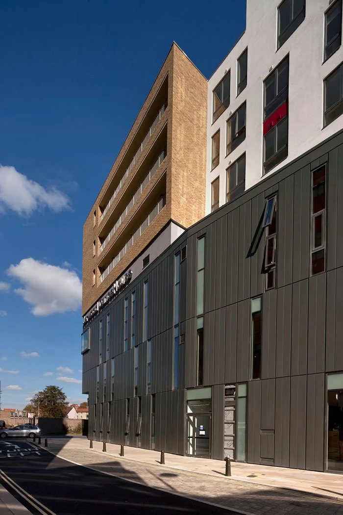 Photo of a modern multi-story building with a black metal exterior on the lower floors and a beige brick upper section, with a clear blue sky and some clouds in the background.