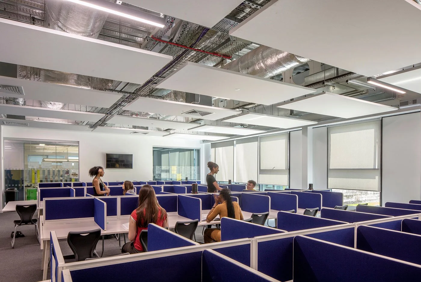 A modern office or classroom space with multiple blue and black chairs and cubicles, several people seated and standing, and a large window with blinds on the right side.