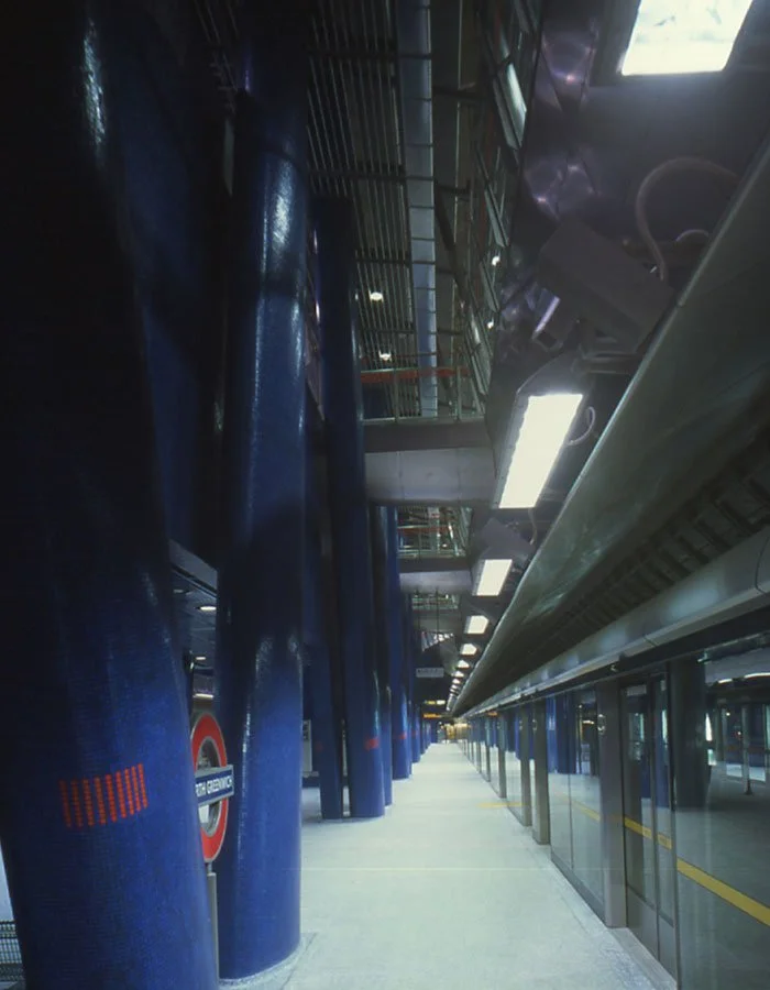 Looking down the platform at an empty North Greenwich underground station platform with blue padded columns, glass walls, and high-tech lighting.