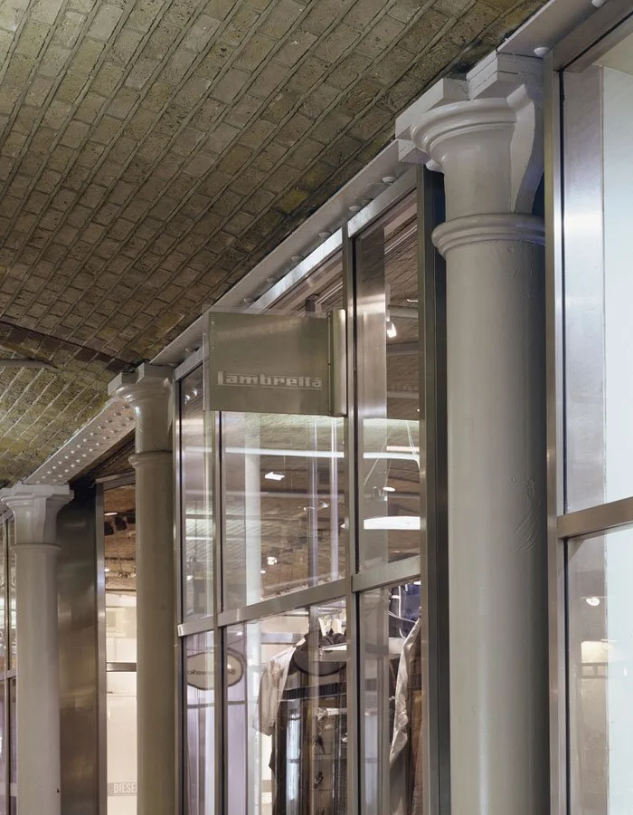 Interior view of a modern side to the Thomas Neal’s Centre commercial building with large glass windows, white columns, and exposed brick ceiling, showing clothing displays inside.