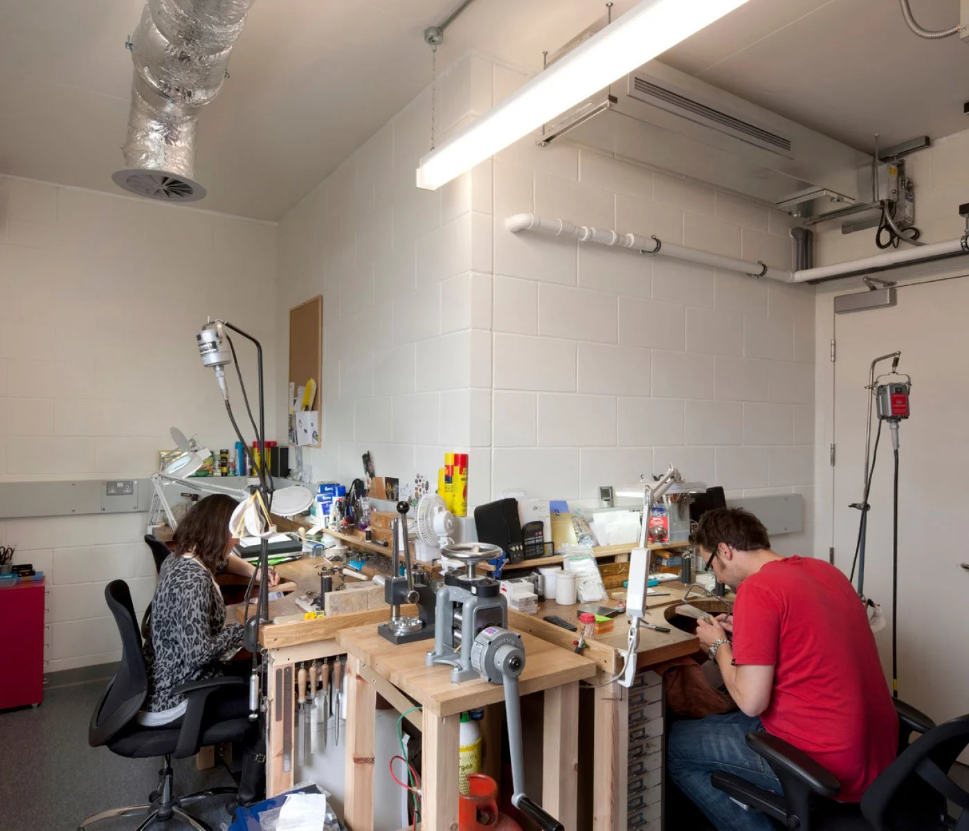 Two people working at a cluttered workbench in a workshop or lab, with tools, paper, and equipment. One person, a woman, is on the left, and a man, wearing a red shirt, is on the right, both focused on their tasks.