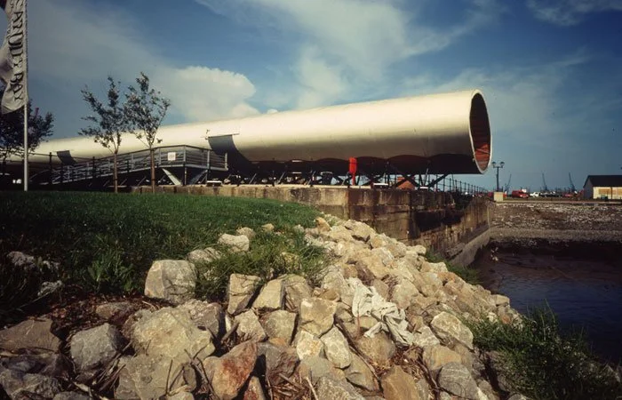 Another view of the 70 metre long squashed tube of elliptical steel rings that is the Visitor Centre extends over the water, with rocks of the doc bay in the main foreground.