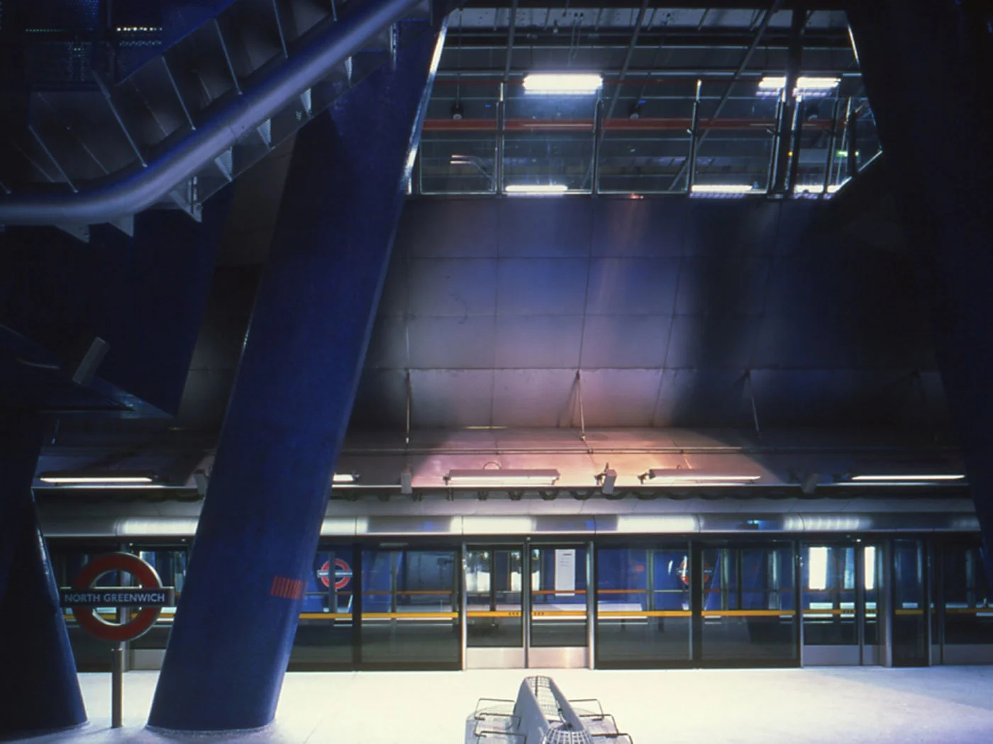 The internal of the North Greenwich underground station with the panel of glass wall and security doors lining the platform edge, with modern architecture and panels in the distance and the large sloping blue columns in the foreground.