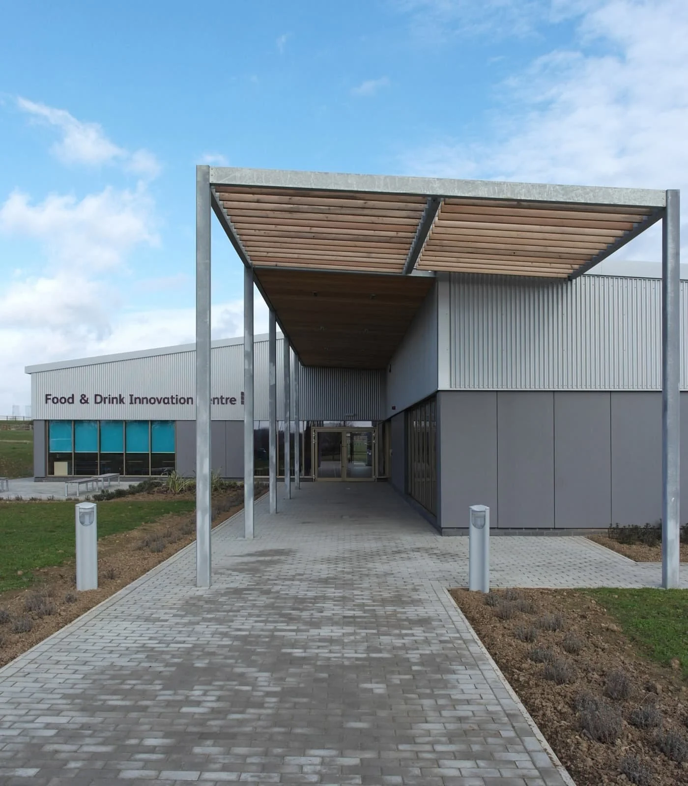 Exterior view of a modern building labeled 'Food & Drink Innovation Centre,' with a paved walkway leading to glass doors, metal and wood architectural features, and a partly cloudy sky overhead.
