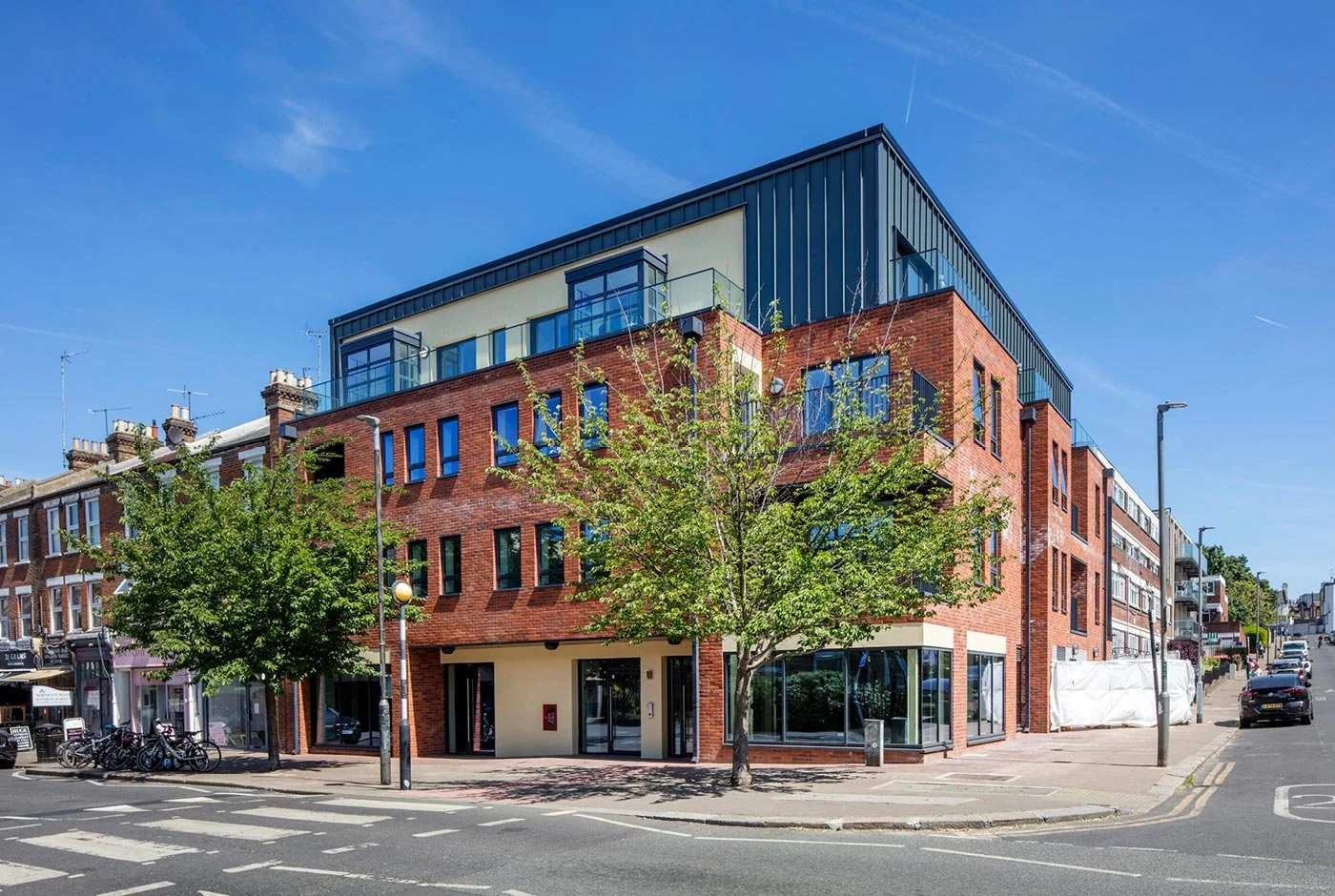 Modern multi-story building with brick and black metal exterior, large glass windows, and greenery in an urban street scene under a blue sky.