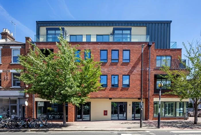 Modern multi-story brick building with large glass windows and balconies, trees in front, bicycles parked to the side, and a clear blue sky.