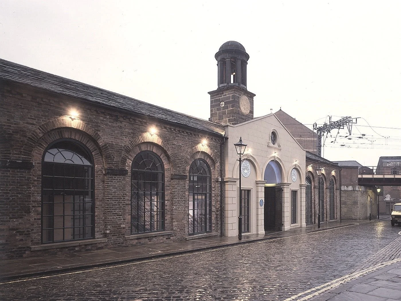 A cobblestone street lined with historic brick and stone building that are the external to White Cloth Hall in Leeds, with street lamps and a cloudy sky overhead.