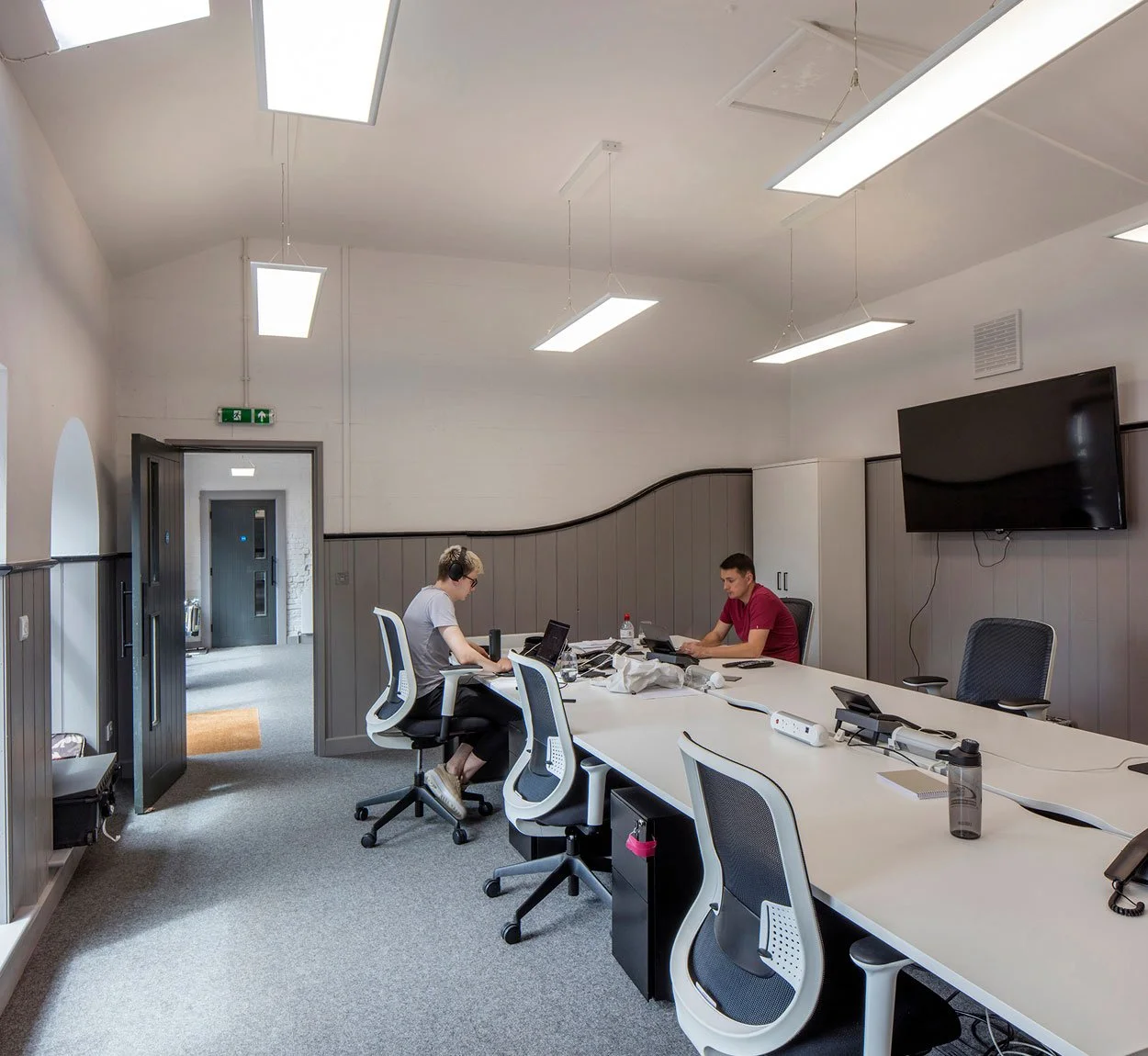 Modern conference room with a long table, two people working on laptops, large wall-mounted TV, gray and white decor, ceiling lights, and open door leading to hallway.
